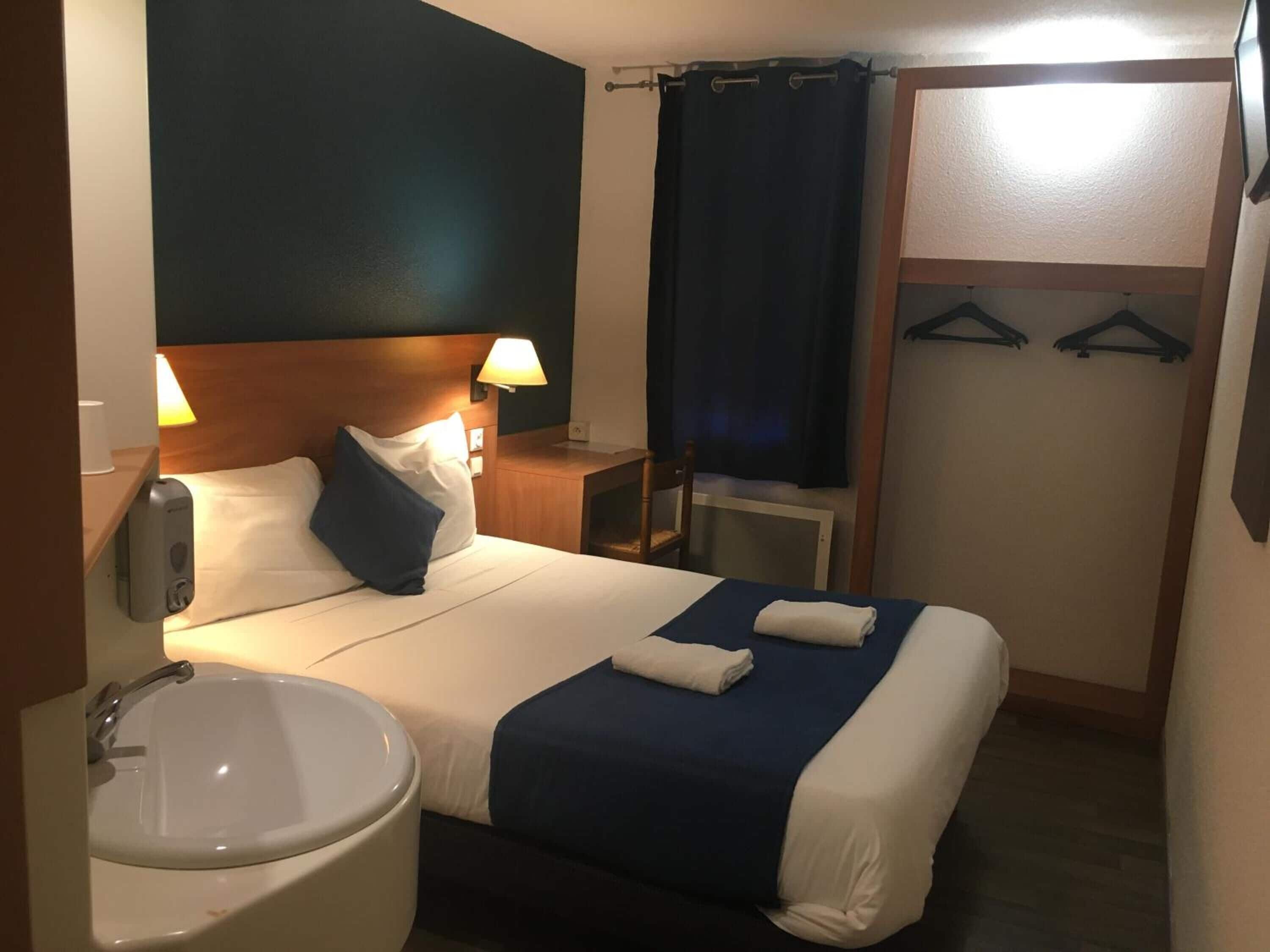 Quarto de hotel com cama, abajures, mesinha, cadeira, pia e guarda-roupa com cabides.