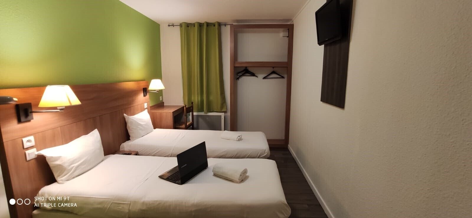 Habitación de hotel con dos camas, un escritorio, una lámpara, una computadora portátil, un televisor y un perchero.
