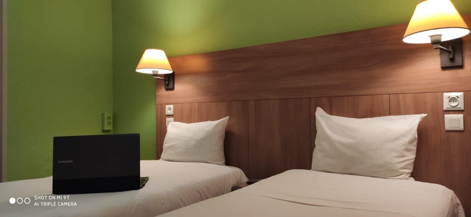 A hotel room with two beds, a wooden headboard, bedside lamps, and a laptop on the left bed.