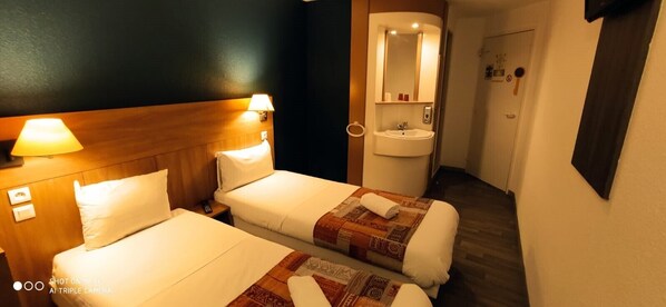 A hotel room with two beds, a wooden headboard, a bedside lamp, a bathroom with a sink and mirror, and a door leading to another room.