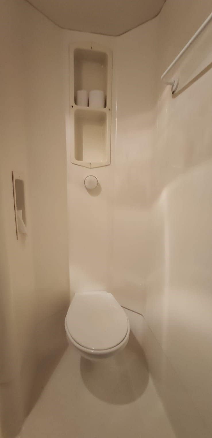 A small, white, enclosed bathroom with a toilet, a shelf with toilet paper, and a towel rack.