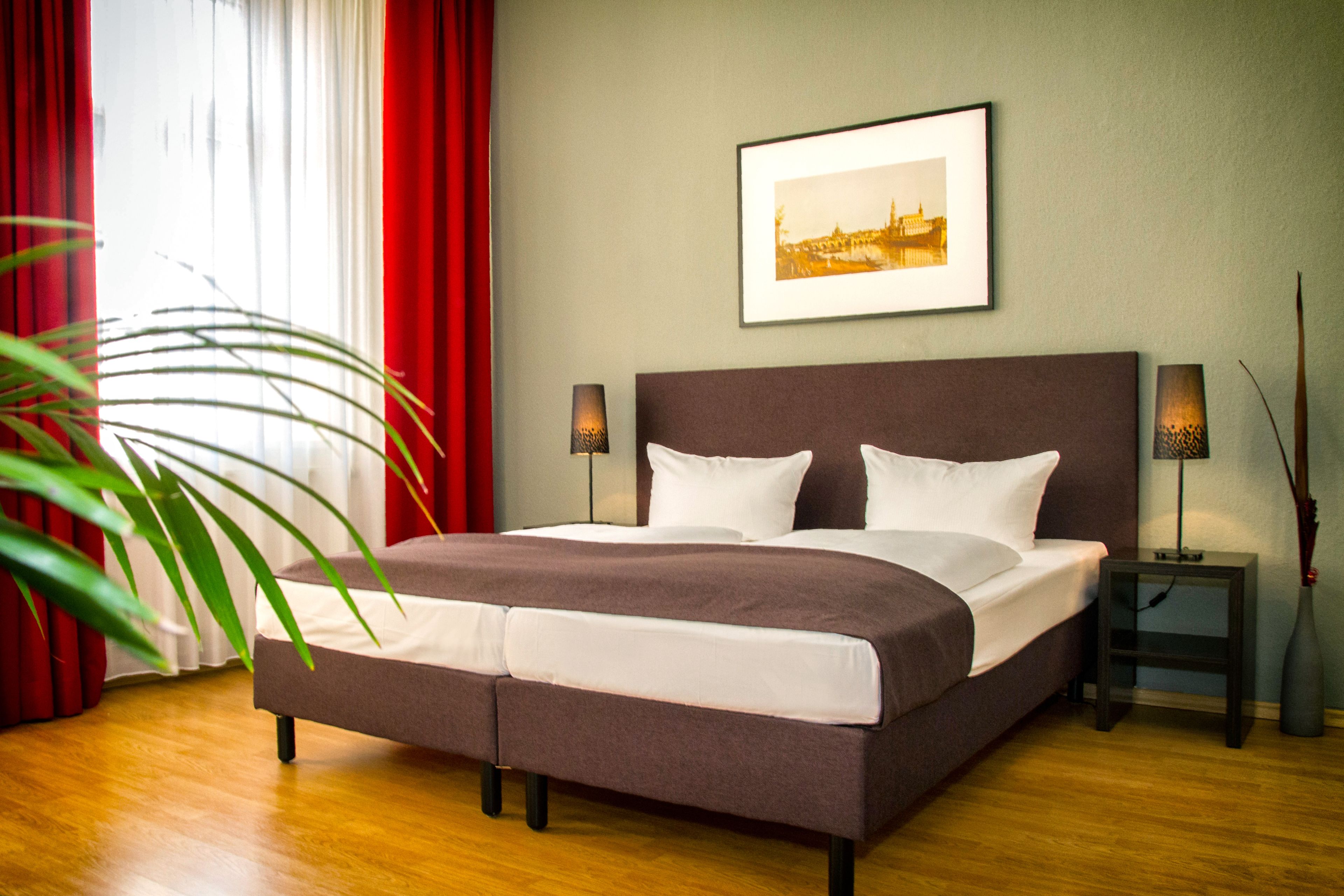A large bed with a headboard, two bedside tables with lamps, a framed picture on the wall, and a potted plant.