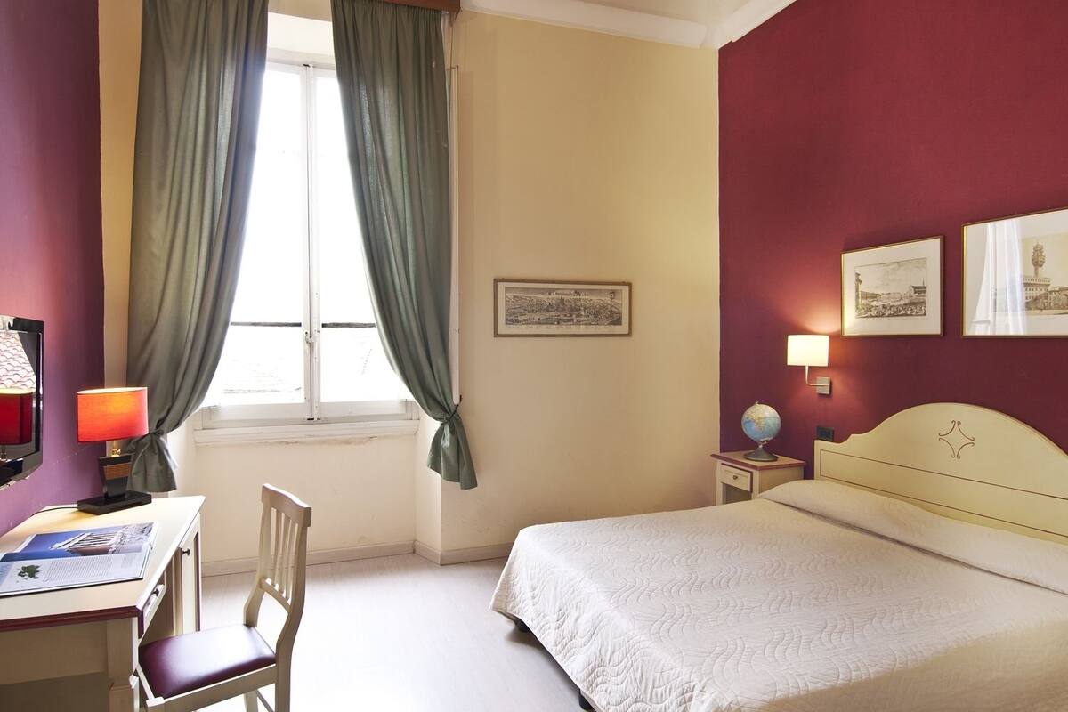 A hotel room with a bed, a desk with a lamp, a chair, a window with curtains, and framed pictures on the wall.