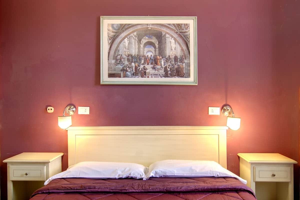 A hotel room with a large bed, two bedside tables, a framed artwork above the bed, and wall-mounted light fixtures.