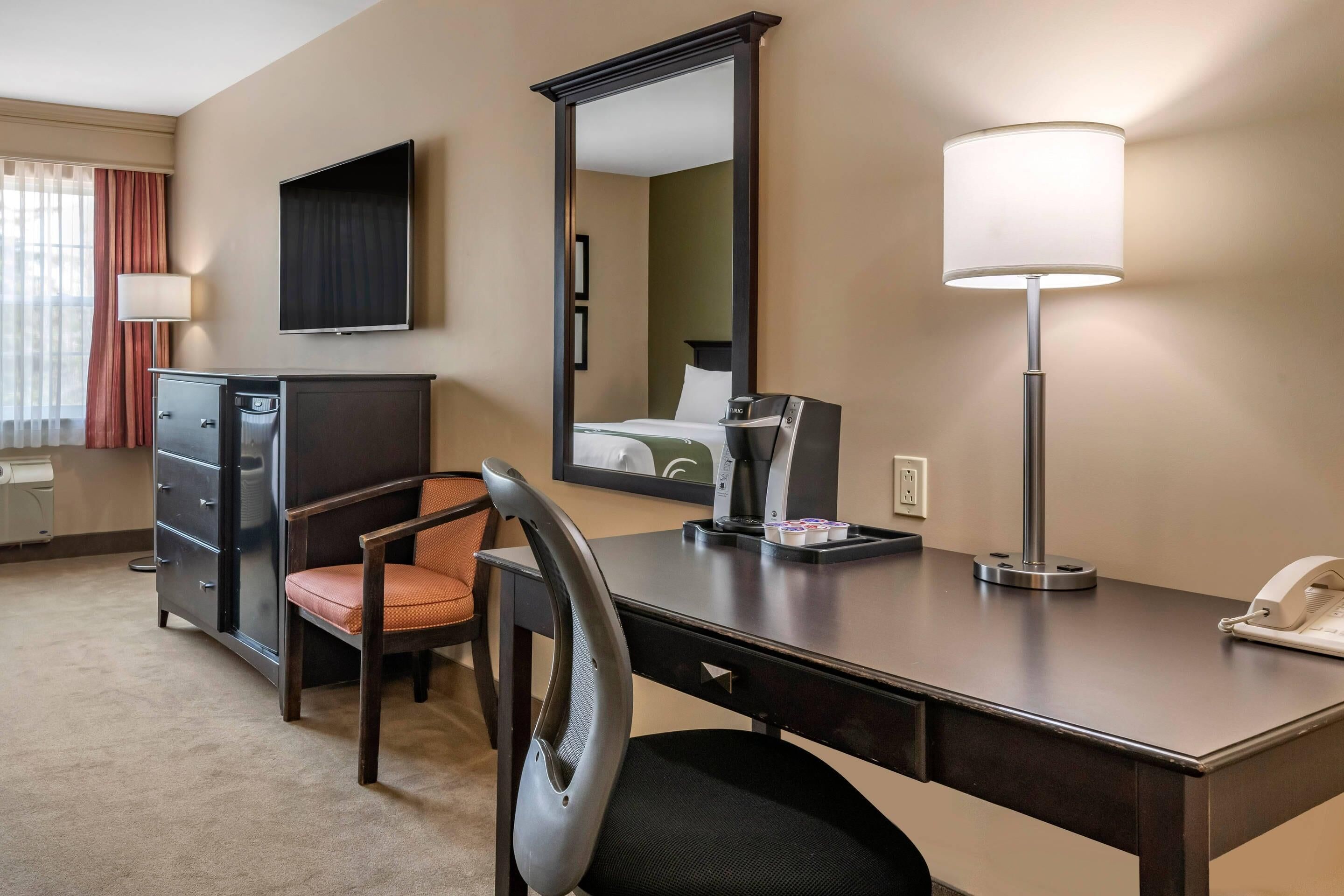 A hotel room with a desk, chair, dresser, and a large mirror.