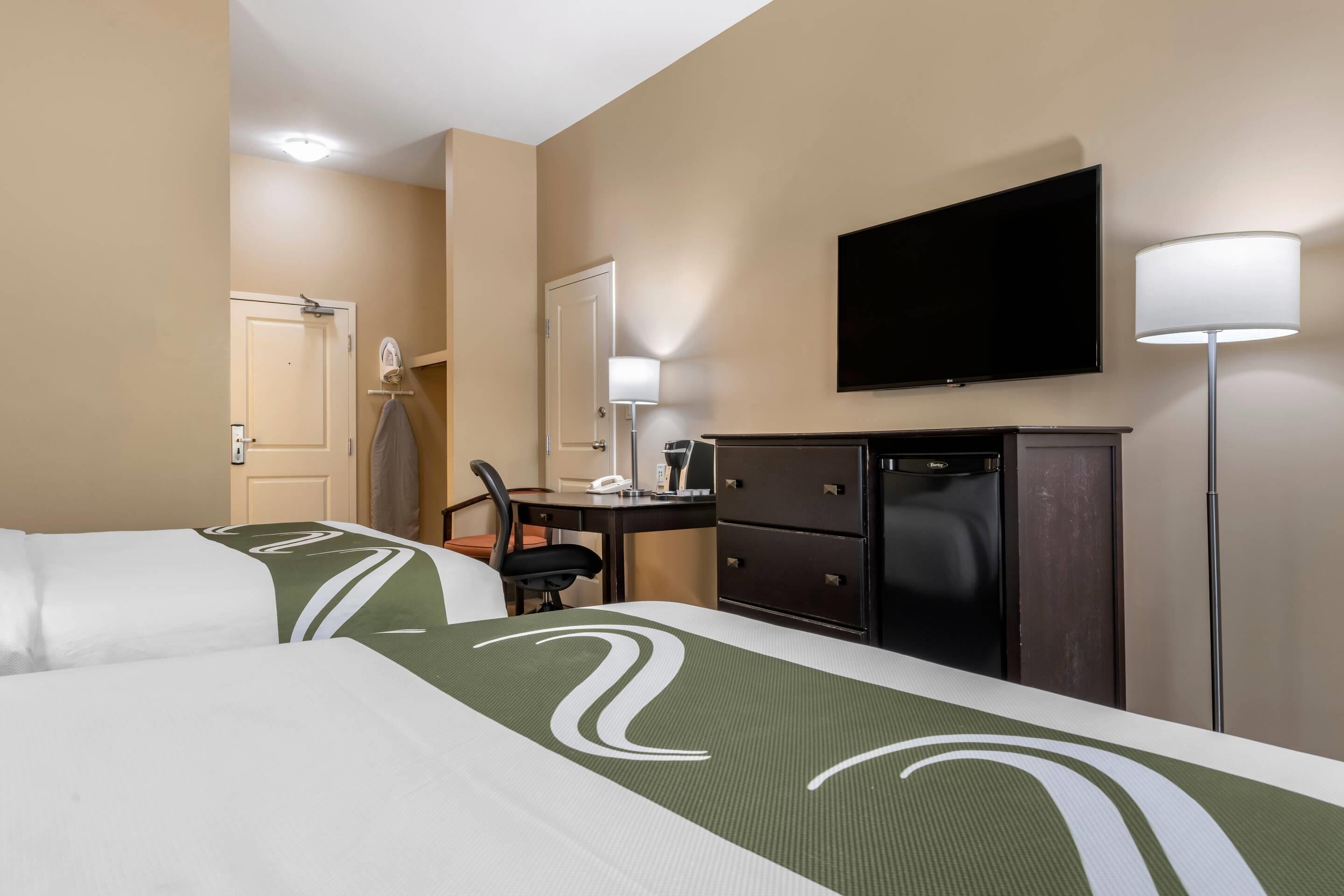 A hotel room with two beds, a desk, a chair, a TV, a lamp, and a minibar.