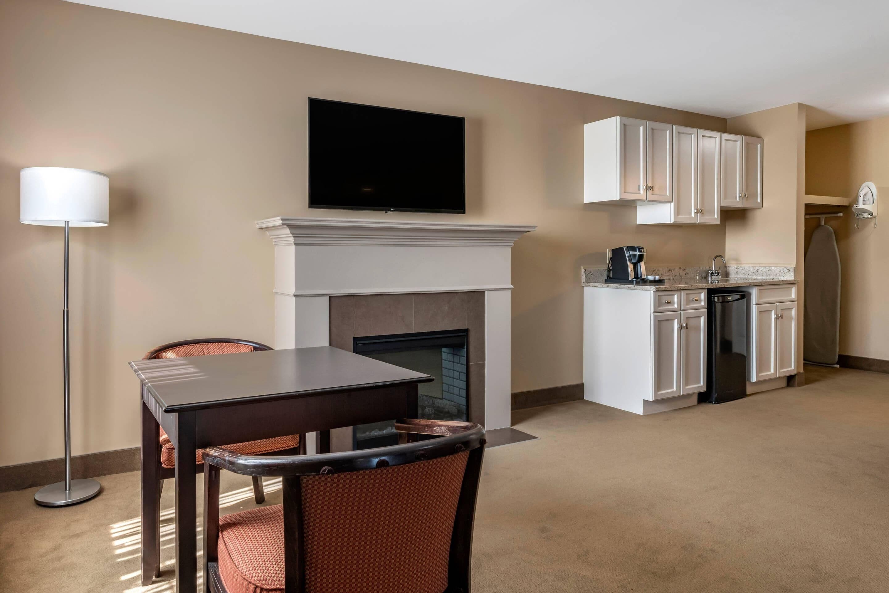 A hotel room with a dining area, a fireplace, a flat-screen TV, a kitchen with white cabinets, and a standing lamp.