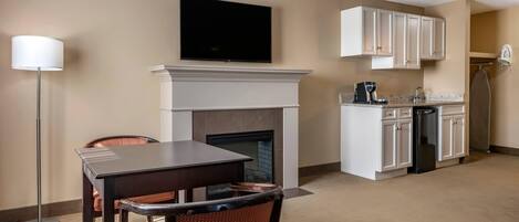 A hotel room with a dining area, a fireplace, a flat-screen TV, a kitchen with white cabinets, and a standing lamp.