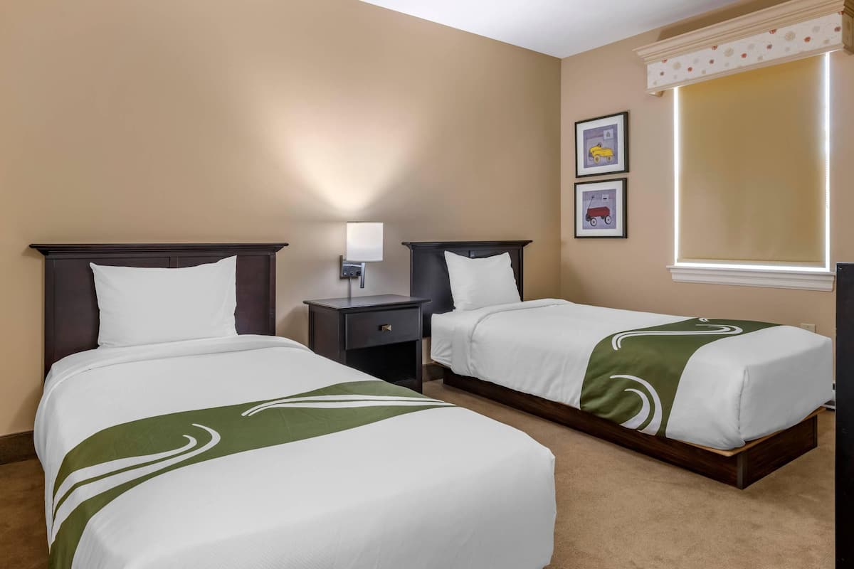 A hotel room with two single beds, a nightstand with a lamp, and a window with blinds.