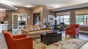 A spacious living room with a beige sofa, a red armchair, and a coffee table. There is a kitchen area in the background with a dining table and chairs. - Quality Inn & Suites Quispamsis (Quispamsis)