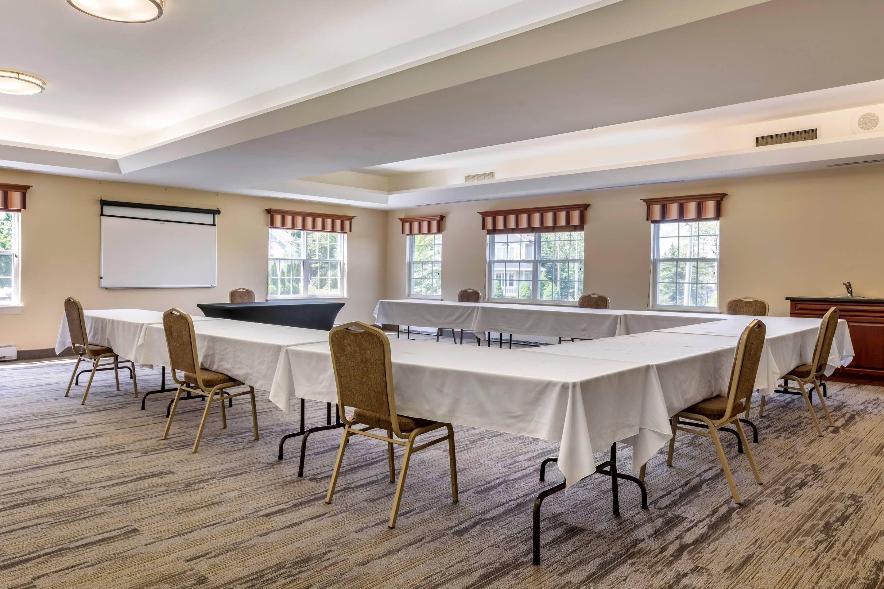 Un salón de conferencias con una mesa larga, sillas, una pantalla de proyector y ventanas con cortinas.