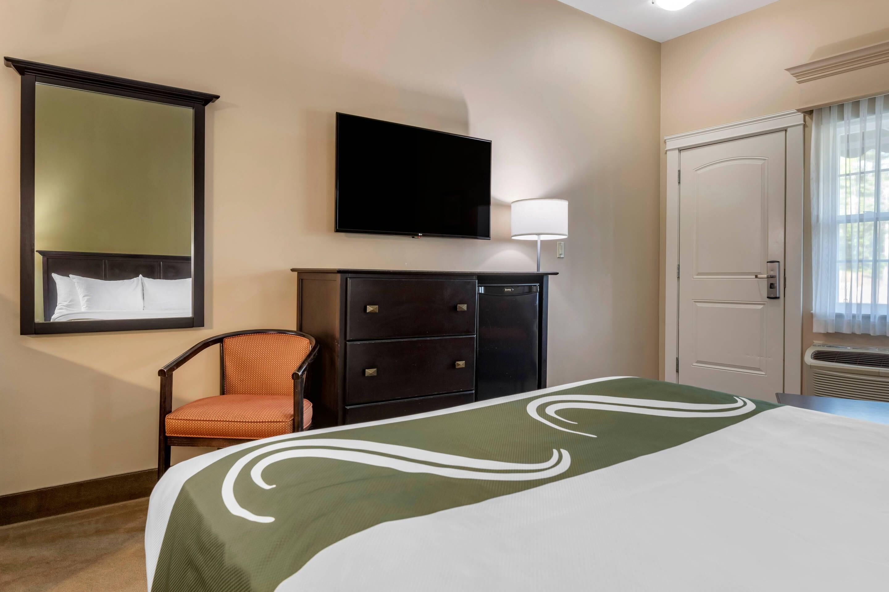 Habitación de hotel con cama, televisor de pantalla plana montado en la pared, una silla, cómoda y puerta con ventana.