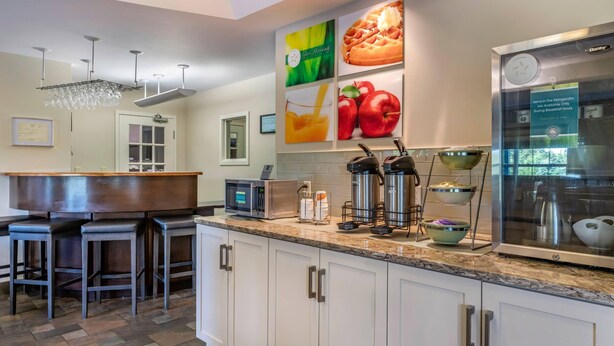 A modern kitchen with white cabinets, a granite countertop, and stainless steel appliances. There are bar stools, a coffee machine, and a refrigerator with a glass door. - Quality Inn & Suites Quispamsis (Quispamsis)