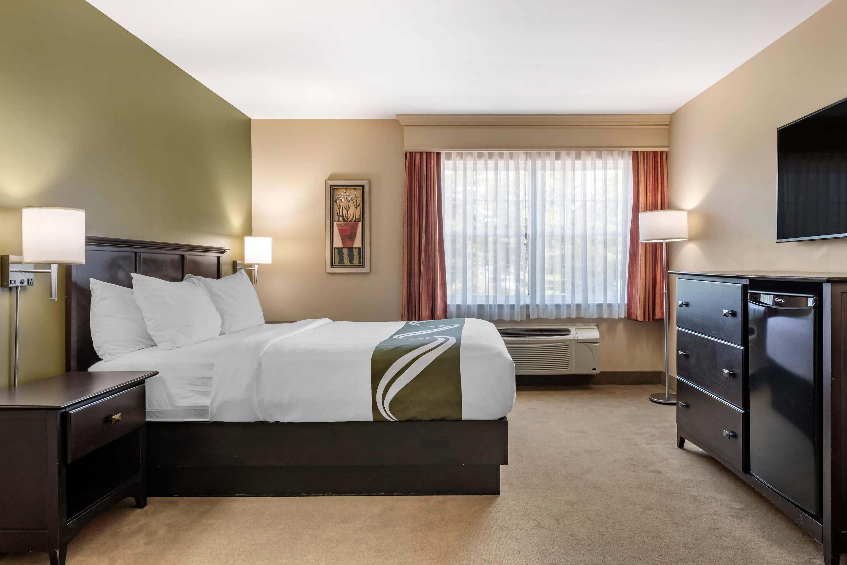 Une chambre d’hôtel comprenant un lit, des tables de chevet, une commode, un téléviseur à écran plat, une fenêtre avec des rideaux et un climatiseur fixé au mur.