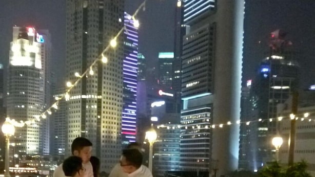 Zona de comedor en la azotea con mesas y sillas, con vistas a un paisaje urbano con rascacielos iluminados.