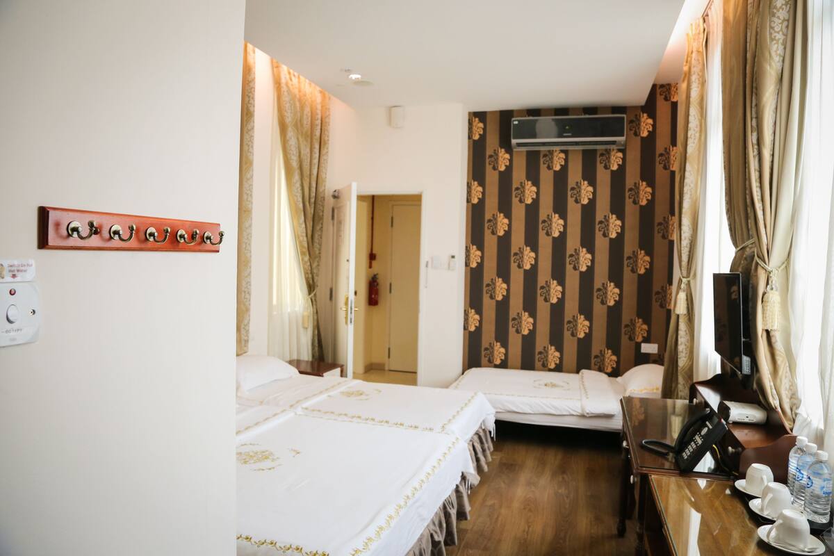 Quarto de hotel com duas camas, uma escrivaninha de madeira com telefone e uma garrafa de água, e um ar condicionado fixado na parede.