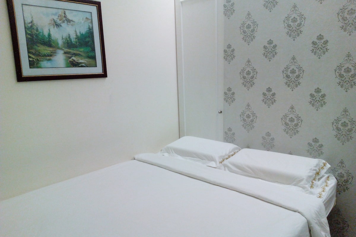 Uma cama de casal com lençóis brancos em quarto com papel de parede estampado e uma pintura de paisagem emoldurada na parede.