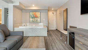 A modern hotel room with a gray sofa, a wooden coffee table, a large mirror, a flat-screen TV, a white bathroom with a large mirror, and a painting on the wall.