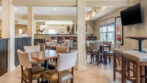 A spacious restaurant with multiple dining tables and chairs, a kitchen area in the background, and a flat-screen TV mounted on the wall.