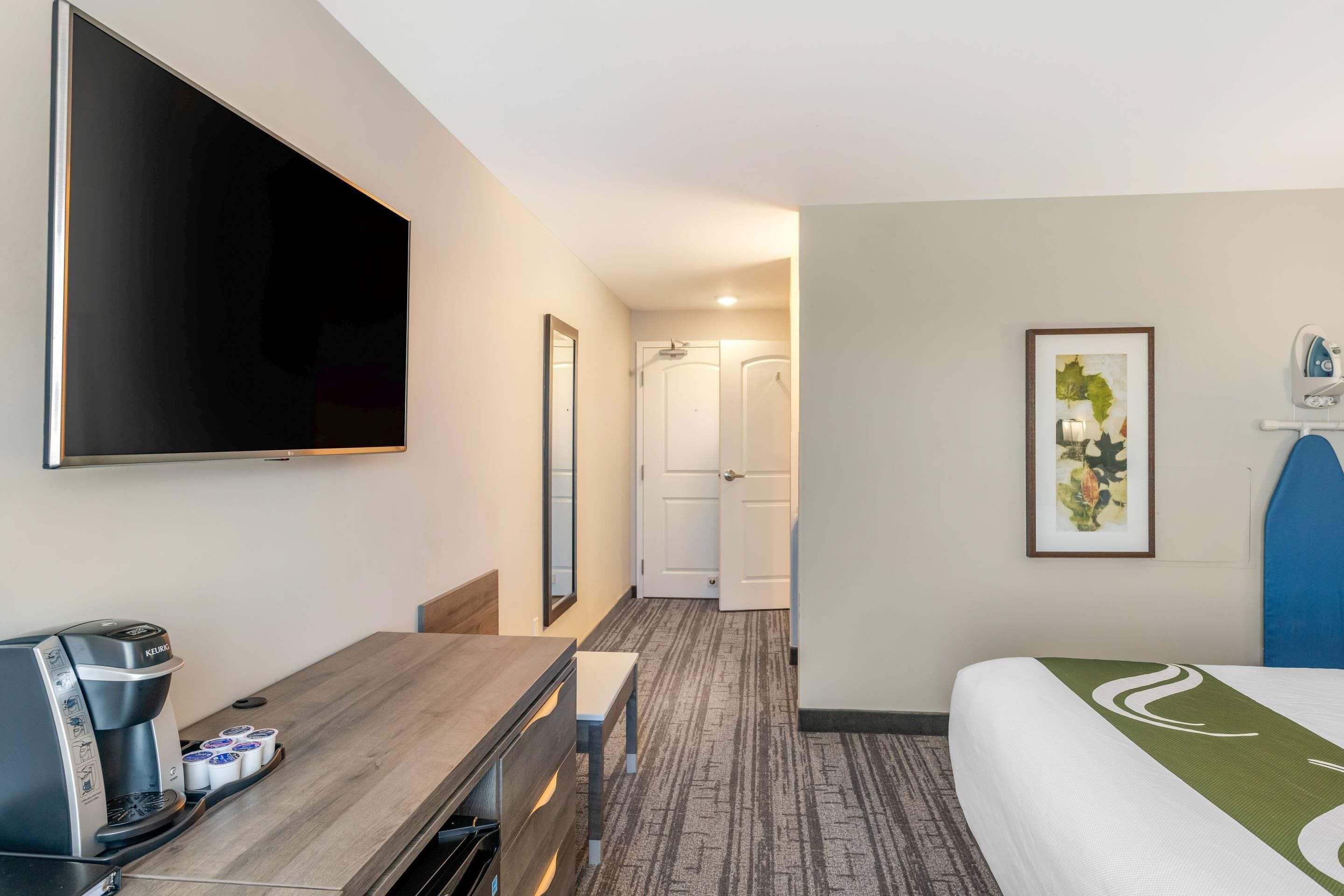 Camera d'albergo con televisione a schermo piatto, macchina del caffè, scrivania, sedia, letto con copripiumino verde e bianco e un quadro appeso al muro.