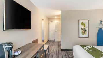 Camera d'albergo con televisione a schermo piatto, macchina del caffè, scrivania, sedia, letto con copripiumino verde e bianco e un quadro appeso al muro.
