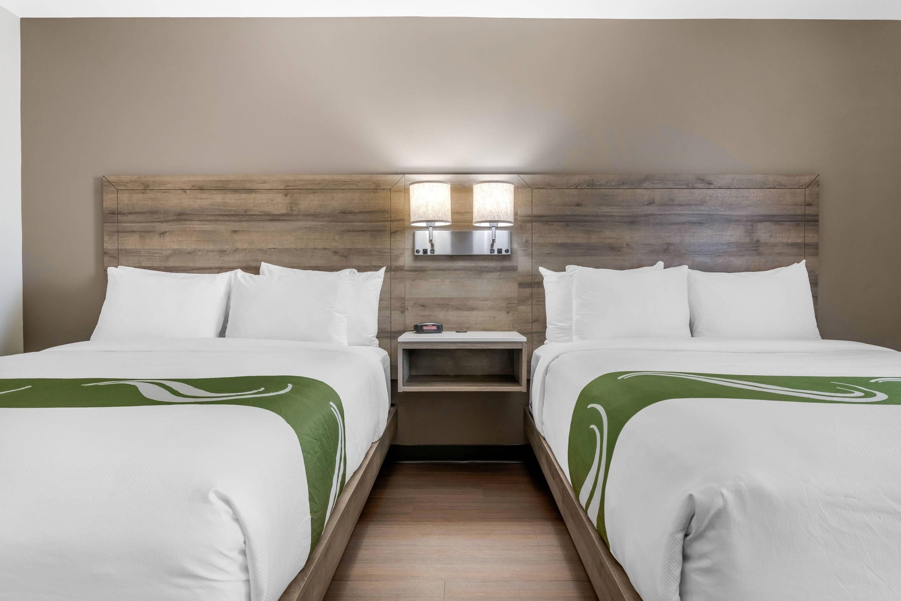 A hotel room with two beds, a wooden headboard, a nightstand, and a wall-mounted light fixture.