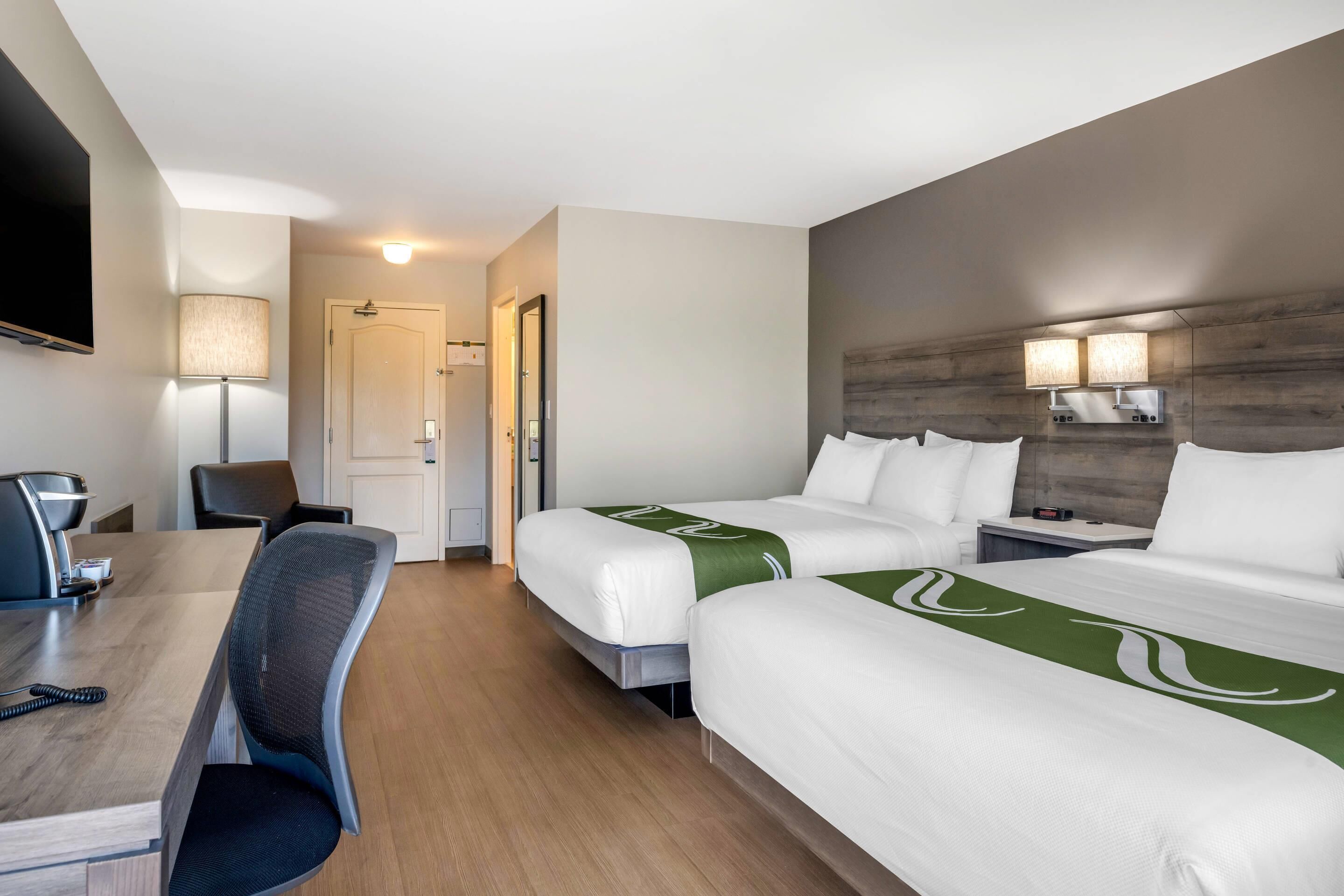 A hotel room with two single beds, a desk with a chair, a TV, and a lamp.