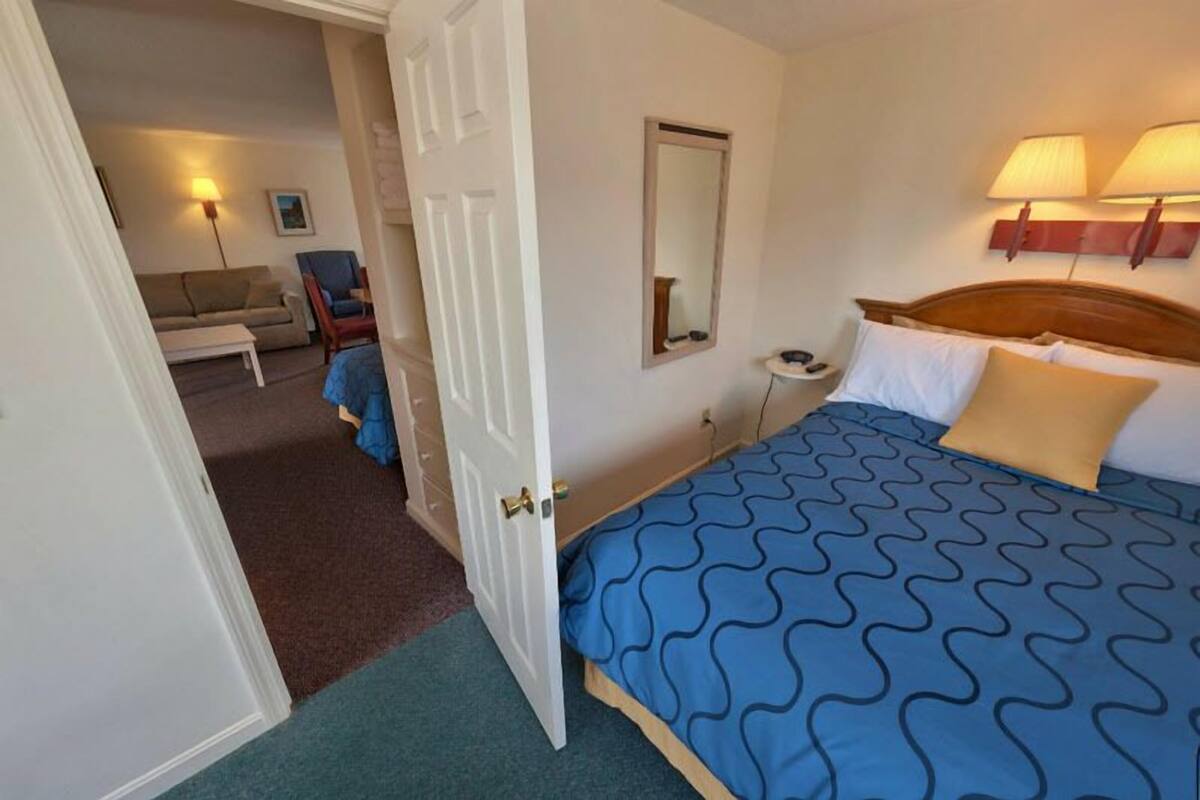 A hotel room with a bed, bedside table, lamp, and a view into the living area.