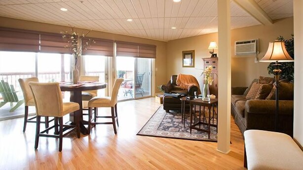 A spacious living area with a dining table, chairs, a sofa, and a coffee table. There is a large window providing natural light and a view of the outdoors. - Sands by the Sea Motel (York Beach)