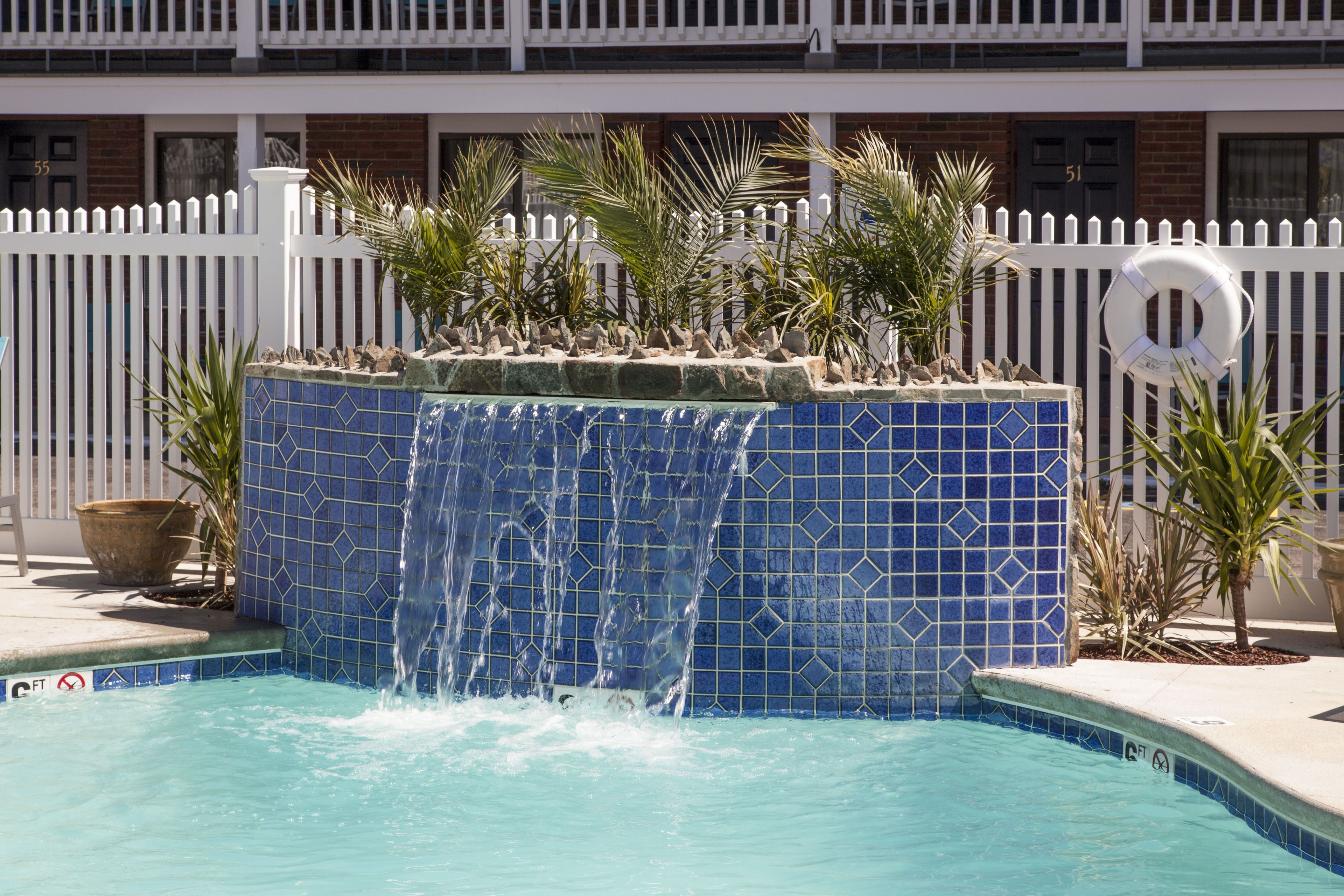 Ein Swimmingpool mit einem blauen Wasserfall aus Fliesen, umgeben von einem weißen Zaun und Palmen.