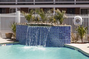 Ein Swimmingpool mit einem blauen Wasserfall aus Fliesen, umgeben von einem weißen Zaun und Palmen.