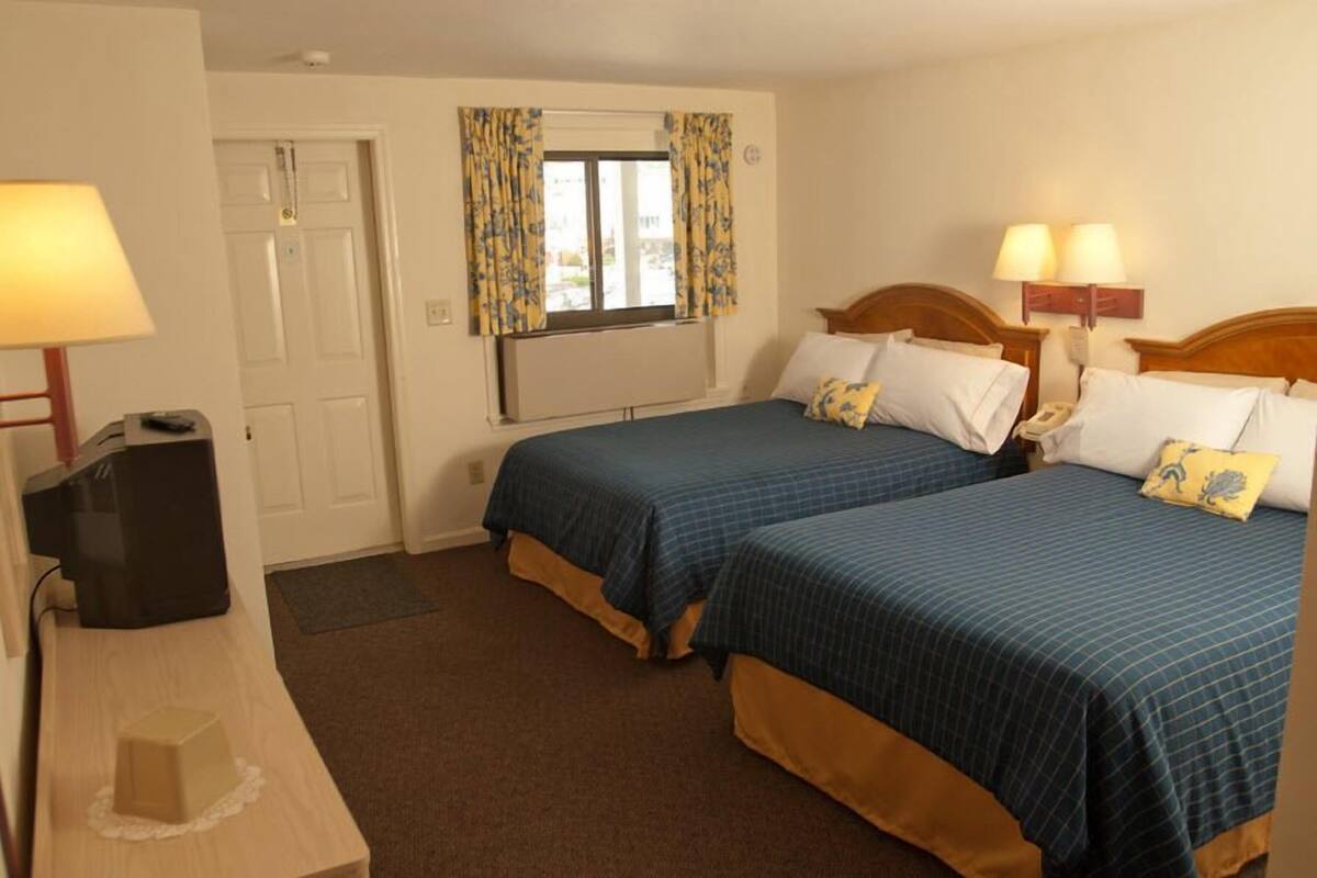A hotel room with two beds, a desk with a lamp, a telephone, and a window with curtains.