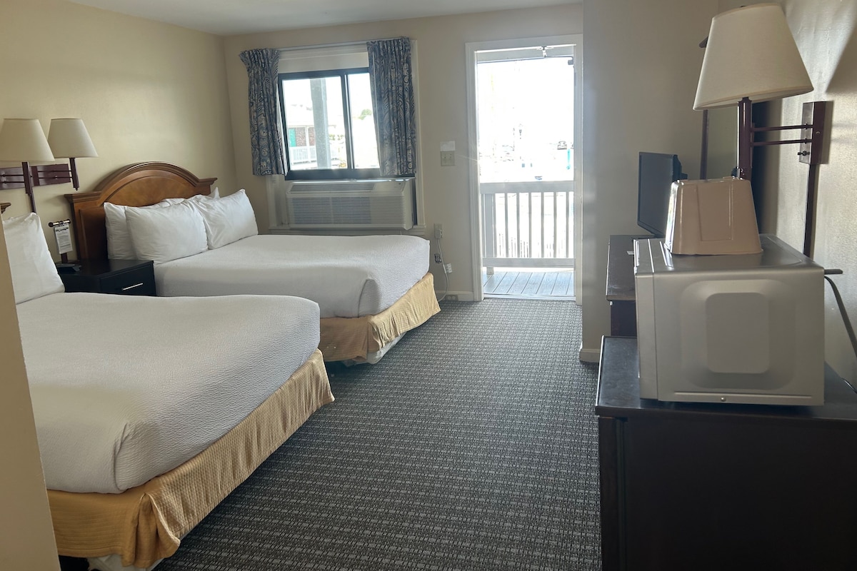 A hotel room with two beds, a desk with a lamp, a microwave, and a window with curtains.