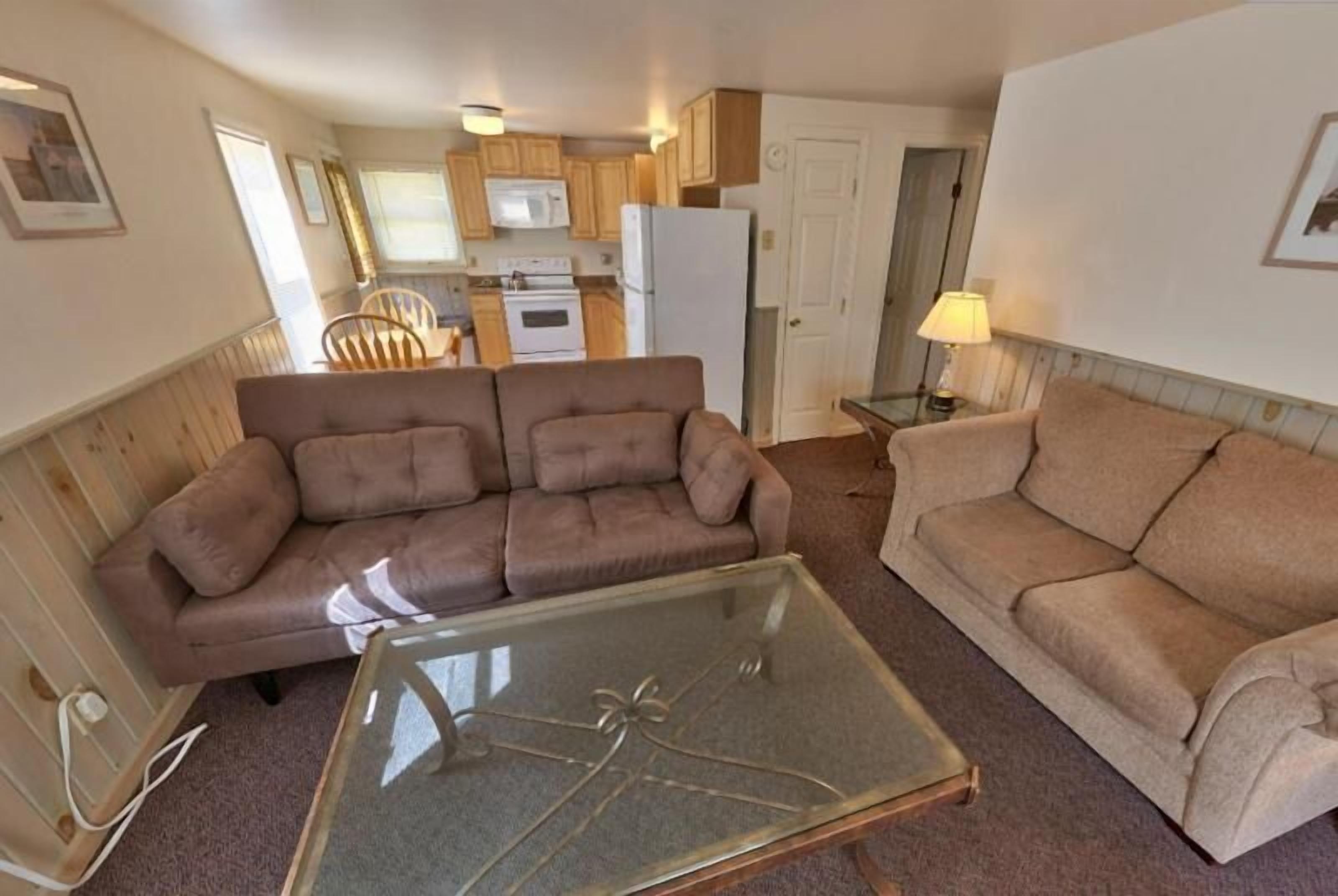 A compact living space with a sofa, a glass-top coffee table, and a kitchenette in the background.