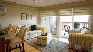 A living room with a sofa, armchairs, a coffee table, and a dining table. There are two framed pictures on the wall, a window with blinds, and a view of the outside balcony. - Sands by the Sea Motel (York Beach)