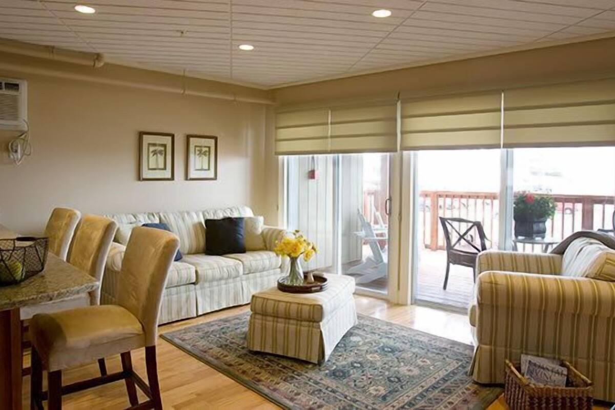 A living room with a sofa, armchairs, a coffee table, and a dining table. There are two framed pictures on the wall, a window with blinds, and a view of the outside balcony.