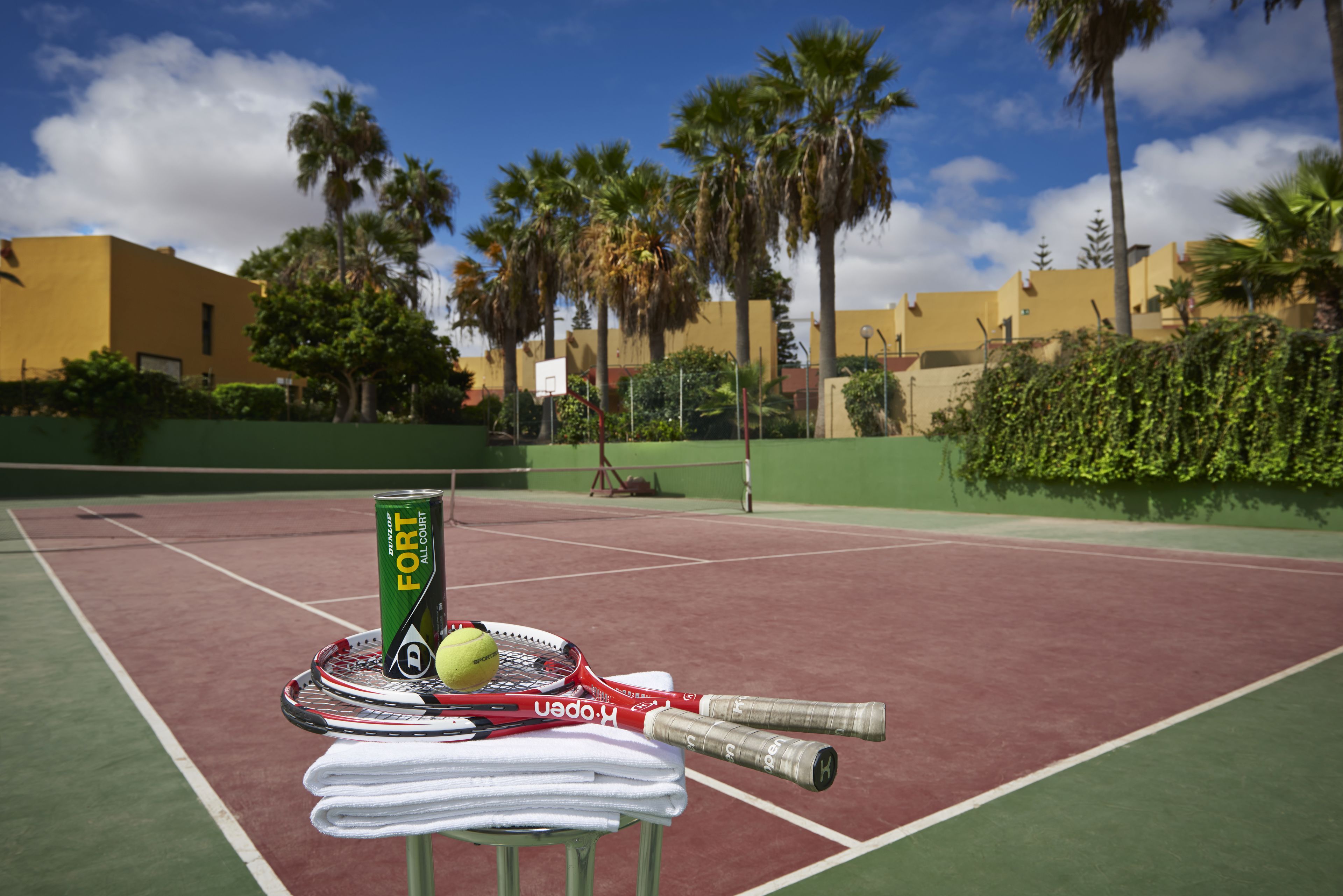 Una cancha de tenis con una mesa dispuesta, raquetas de tenis, una lata de la bebida energética Fortified All Natural, una pelota de tenis y toallas dobladas.