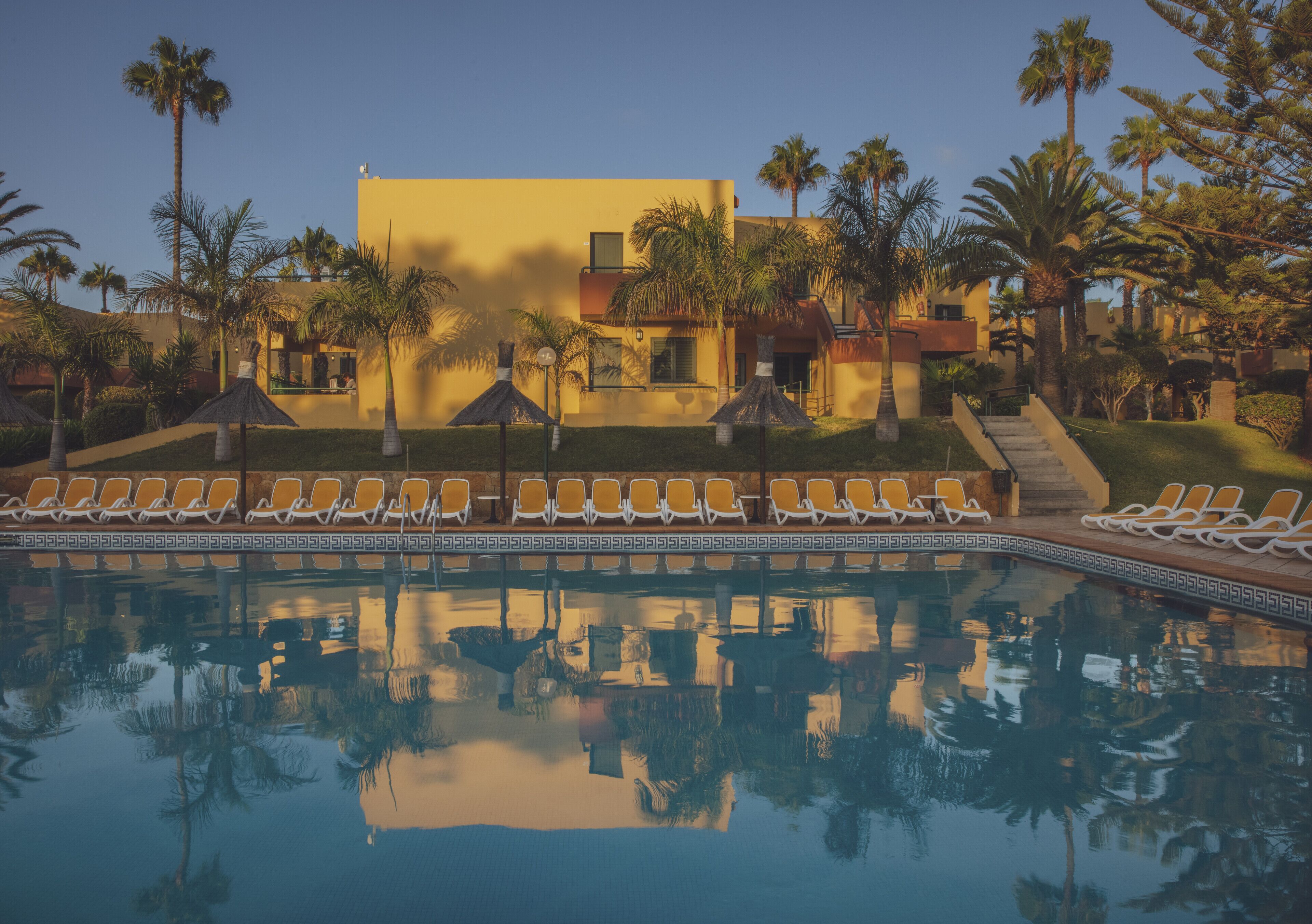 A pool with lounge chairs and palm trees, reflecting the surrounding buildings.