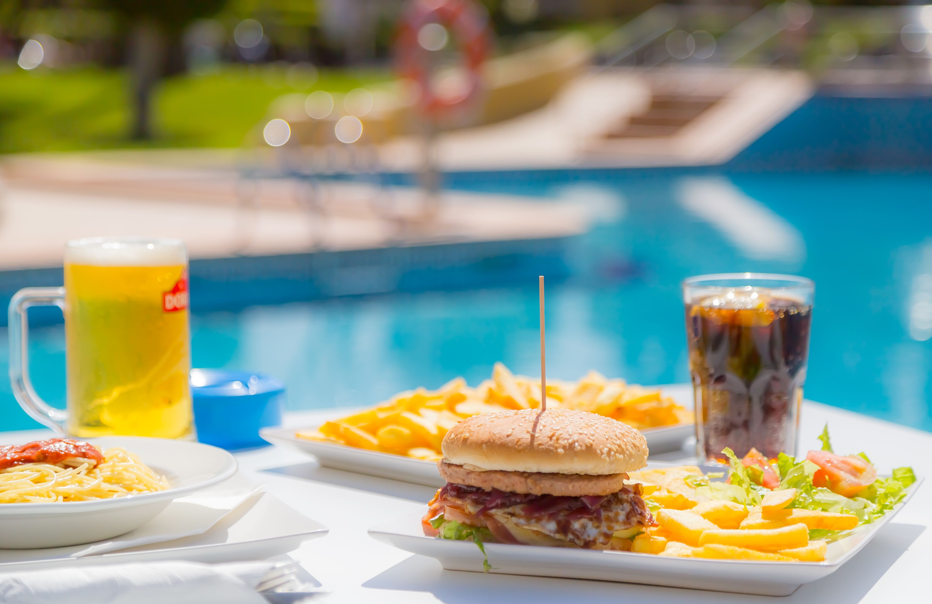 A dining table with a burger, fries, salad, and drinks by a pool.