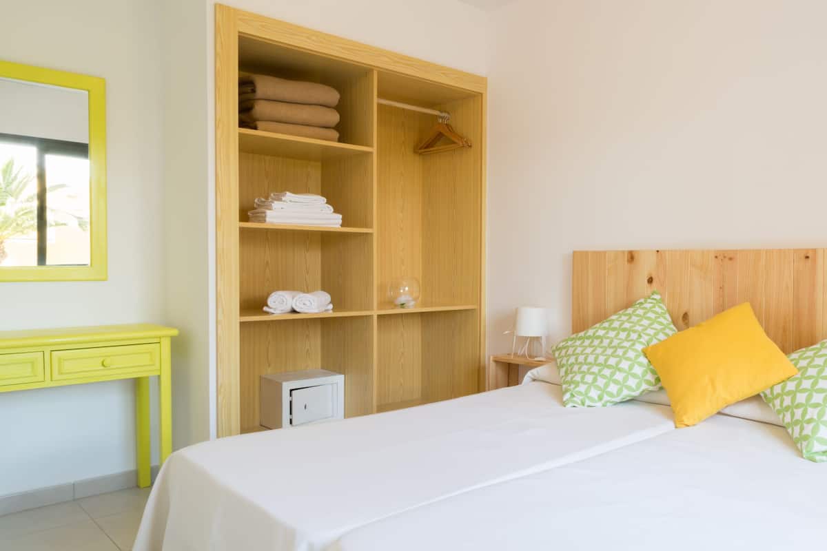 A hotel room with a bed, a wooden wardrobe, a bedside table with a lamp, and a mirror on the wall.