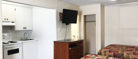 A compact kitchen with white cabinets, a stove, and a sink. There is a wooden dresser with drawers and a flat-screen TV mounted above it. The room has a bed with a colorful quilt.