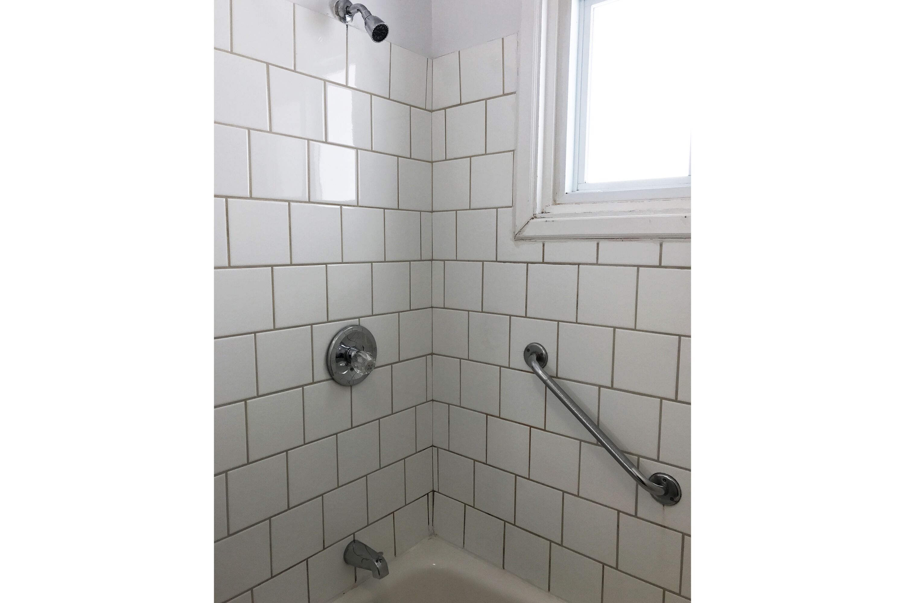 Un baño en esquina con azulejos blancos, un cabezal de ducha, una barra de apoyo y un ventanal.