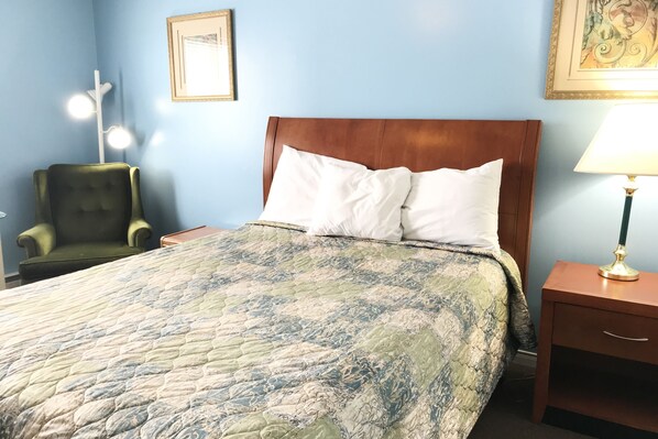 A neatly made bed with a quilted bedspread, a wooden headboard, and two white pillows. There is a green armchair to the left and a nightstand with a lamp to the right. - Bayside Motel (Quinte West)
