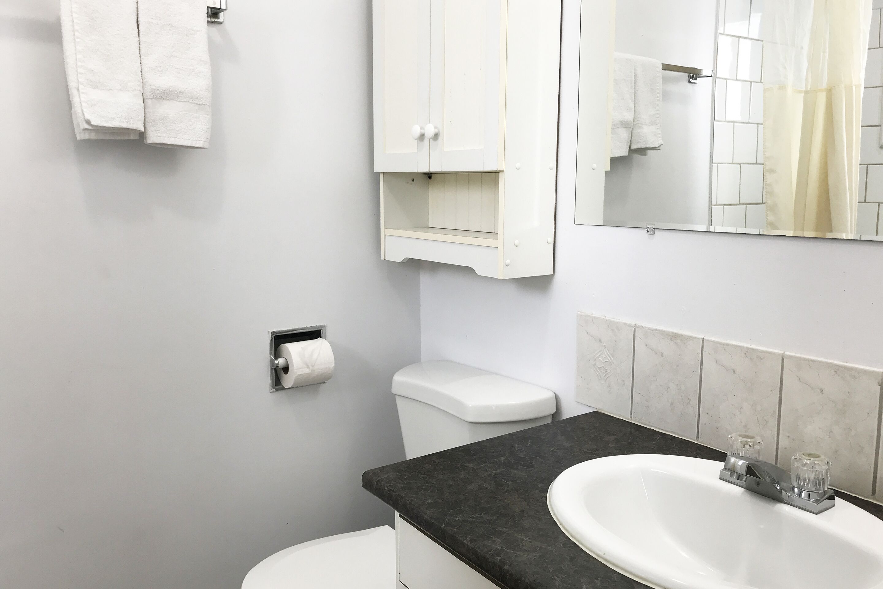 Un baño con inodoro blanco, lavamanos negro sobre encimera y un armario blanco, además de un espejo.