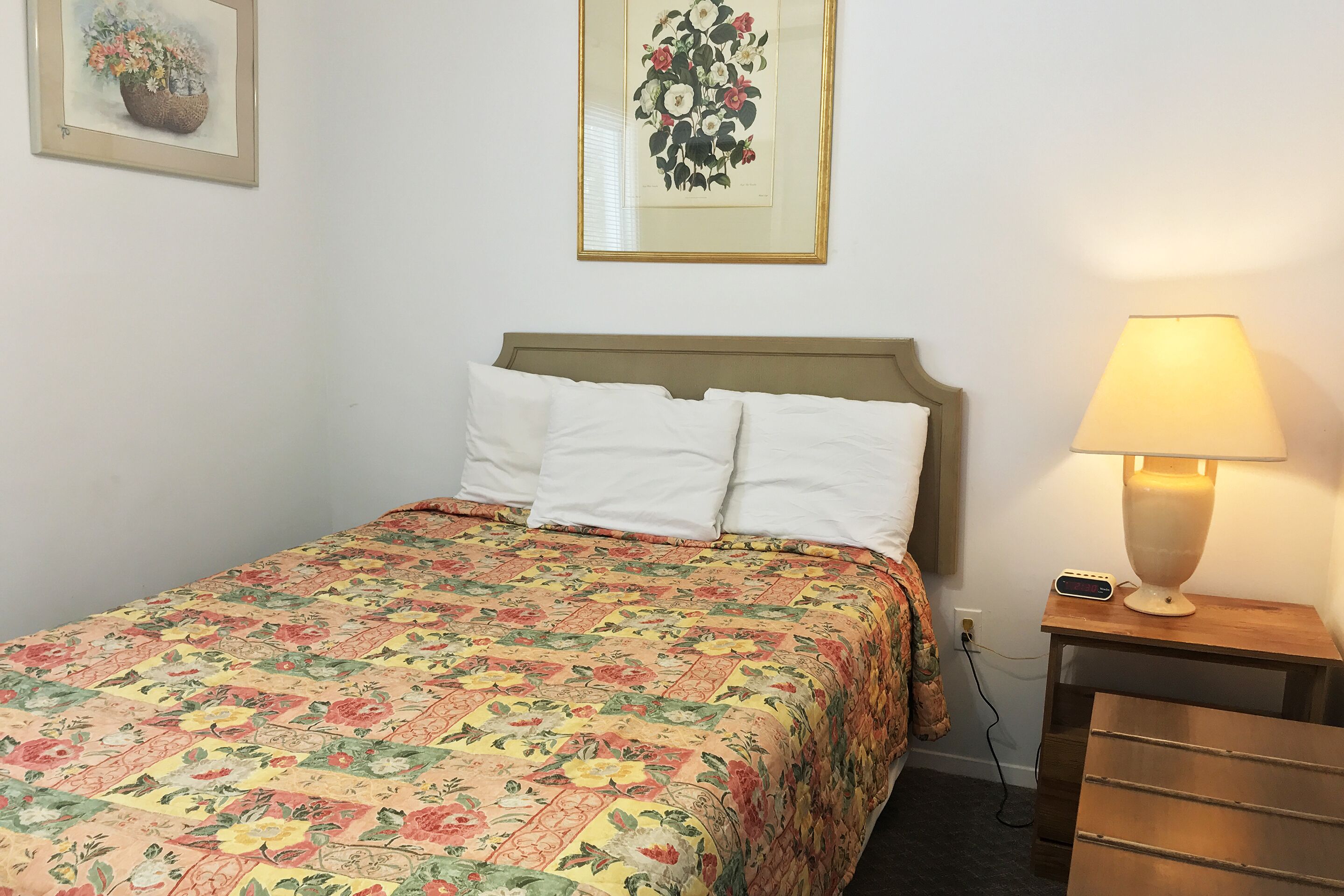 Una cama bien hecha con un edredón floral, una lámpara de noche y una mesita con lámpara.