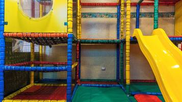 Zona de juegos interior con un tobogán amarillo, estructuras de escalada con red y un ventanal circular.