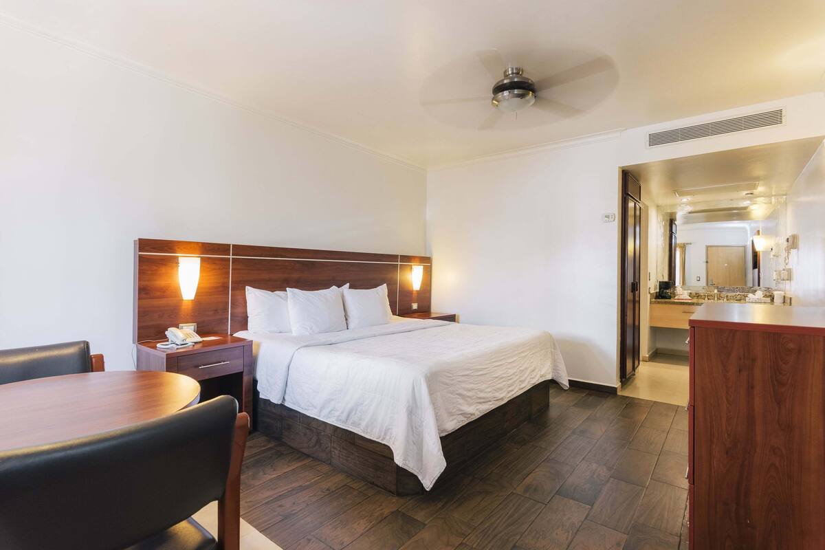 A hotel room with a wooden headboard, a bed with white linens, a bedside table with a lamp, a wooden desk, and a chair.