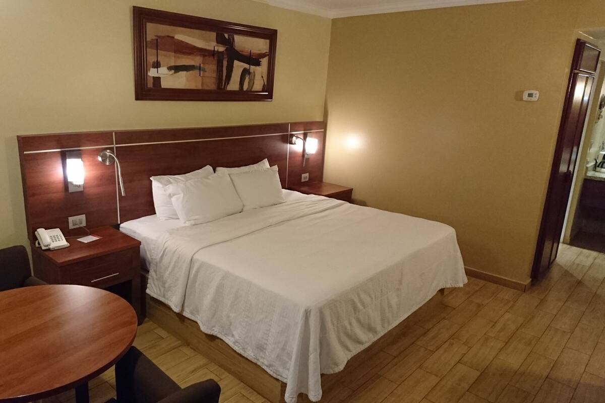 A hotel room with a bed, bedside table, lamp, chair, and a painting on the wall.