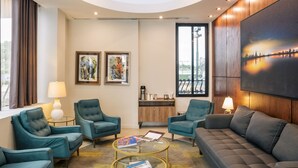 Une chambre d’hôtel moderne avec des fauteuils recouverts d’un tissu turquoise, un canapé gris, une table basse en verre et des œuvres d’art accrochées aux murs.