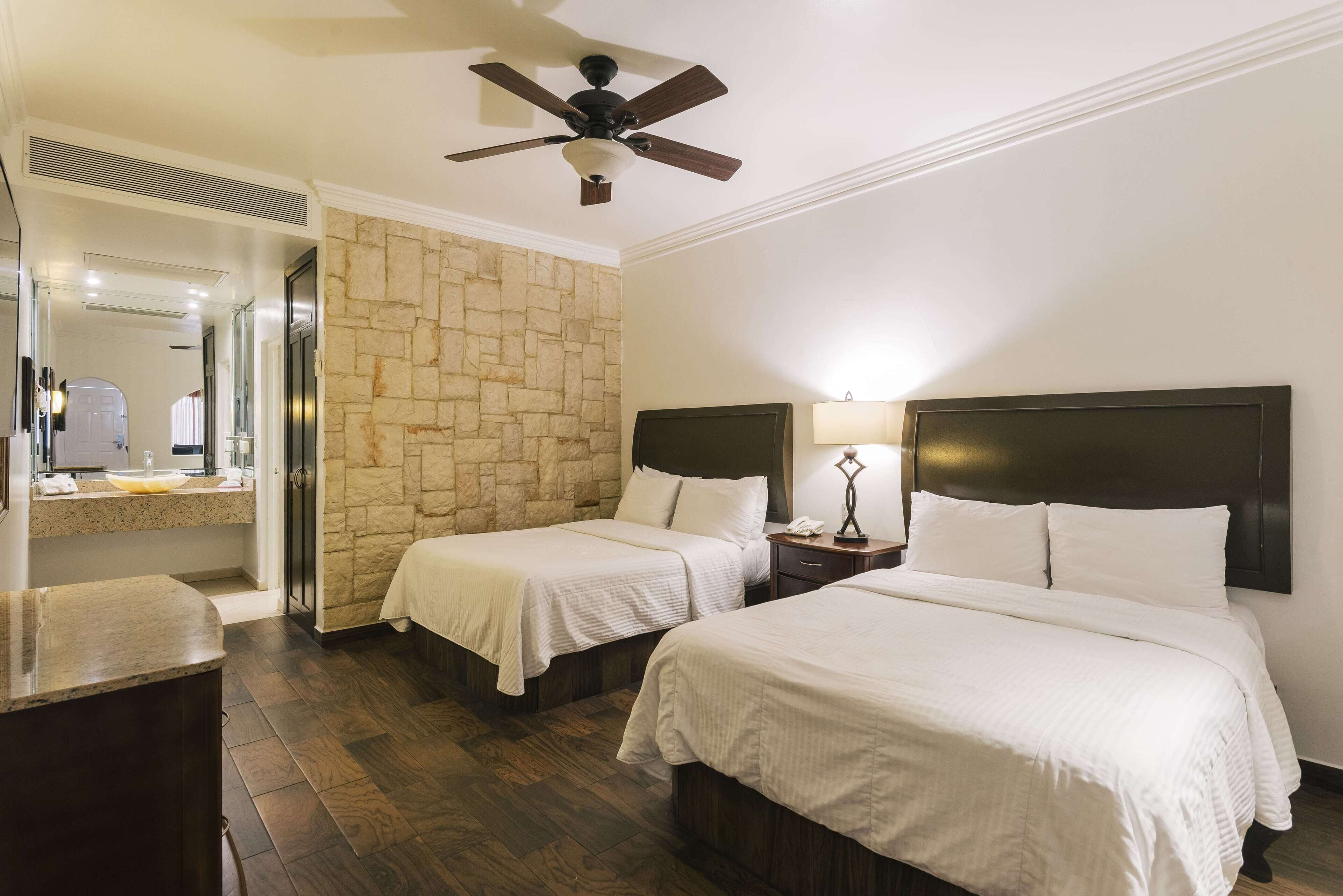 Habitación de hotel con dos camas, una pared de acento de piedra, ventilador de techo, mesita de noche con lámpara y baño con espejo y lavabo.